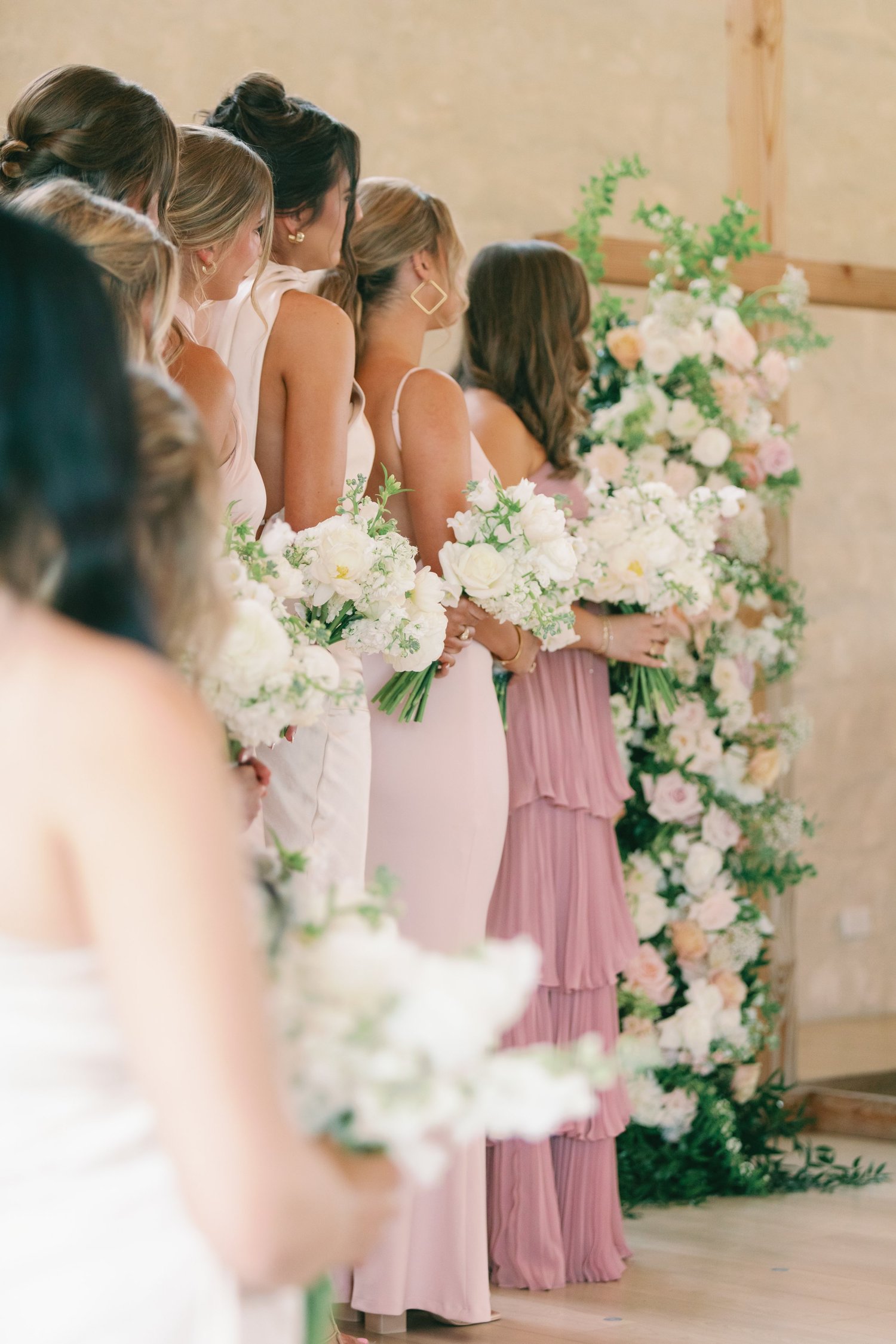 Bridesmaids in blush holding white bouquets by a floral wall